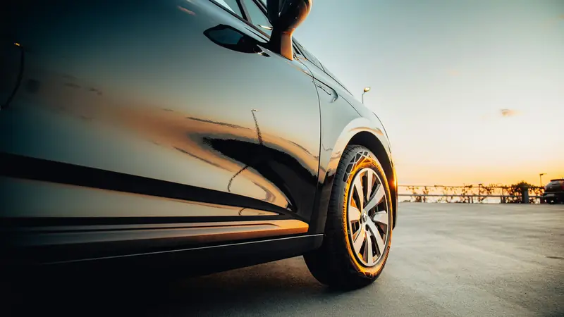 Low-angle view along the glossy flank of a freshly polished dark car, its paint reflecting a golden sunset over the sea